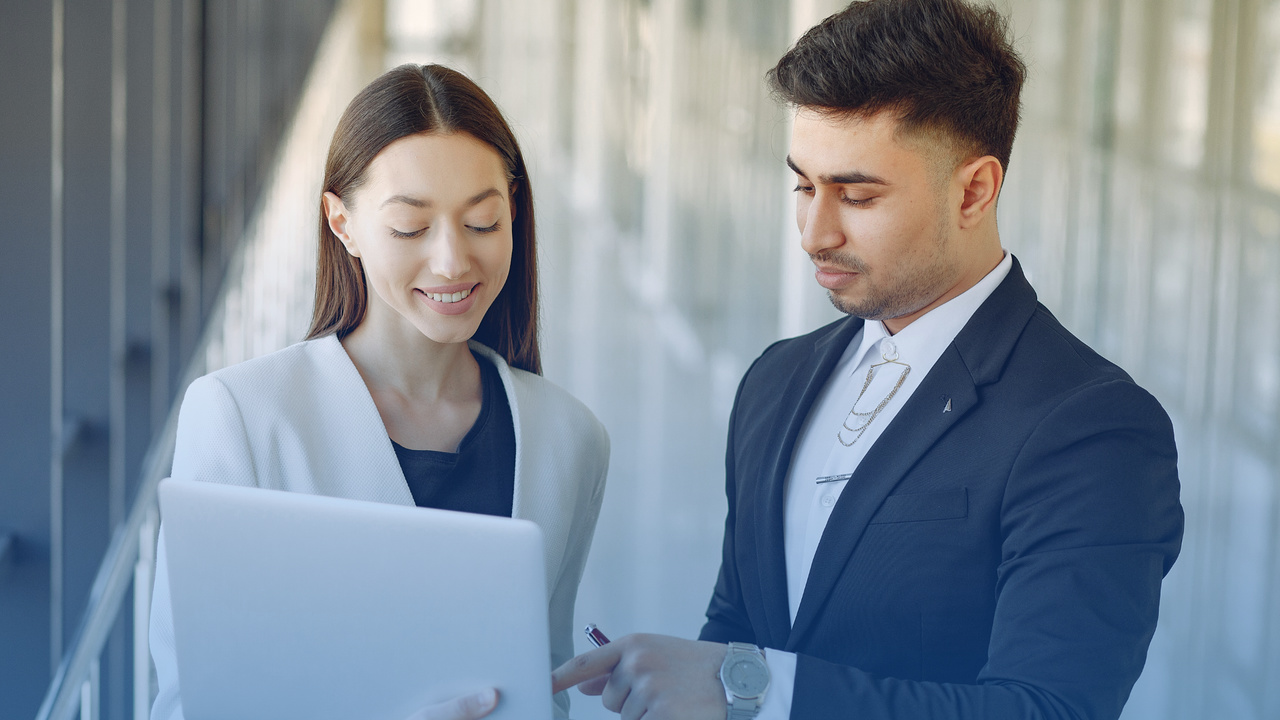 Dois profissionais conversando (homem e mulher) em frente ao notebook. Imagem simboliza que estão acessando os recursos da Área do Cliente do Validador TISS.