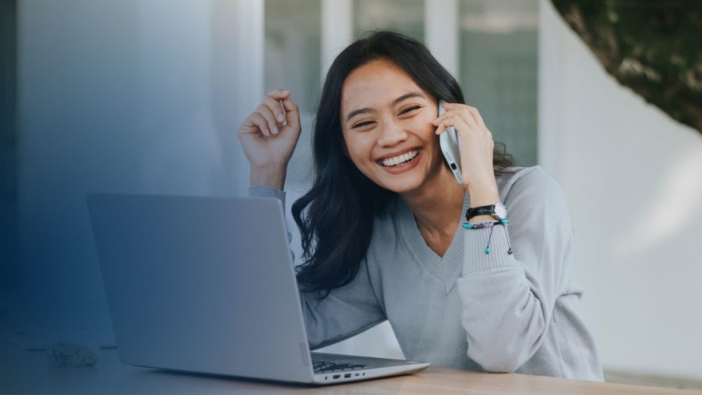 Menina sorrindo falando no celular e de frente para o notebook. Imagem simboliza que ela sabe o que não pode faltar no faturamento das operadoras de saúde.