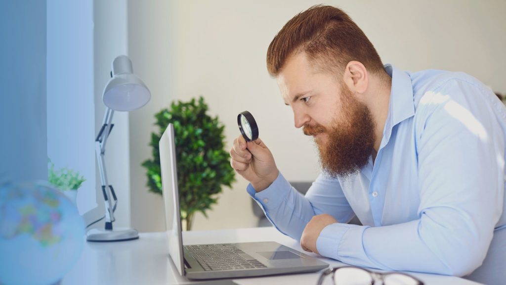 Homem segurando lupa e olhando fixamente para o notebook. Imagem simboliza que ele está fazendo a revisão de guias TISS sem ser automatizada para evitar as glosas.