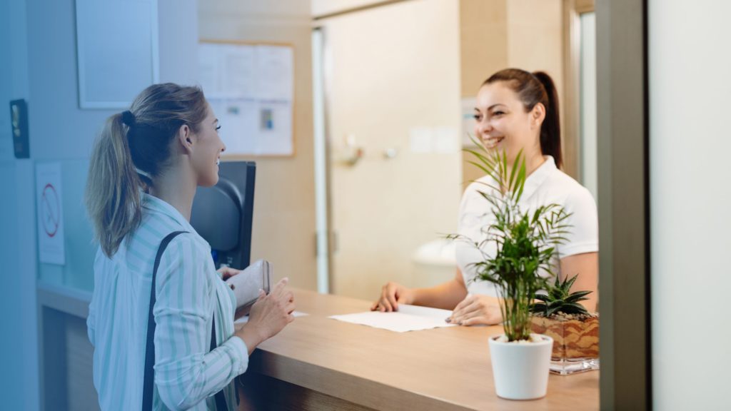 Duas mulheres na recepção de uma clínica médica. A imagem representa a coleta de informações do paciente, antes do atendimento, para evitar a glosa 1006.