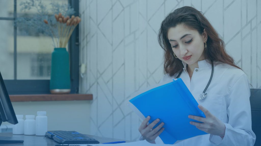 Mulher jovem sentada em sua cadeira dentro de um consultório médico. A imagem representa uma especialista de saúde mensurando a qualidade da assistência na saúde suplementar.