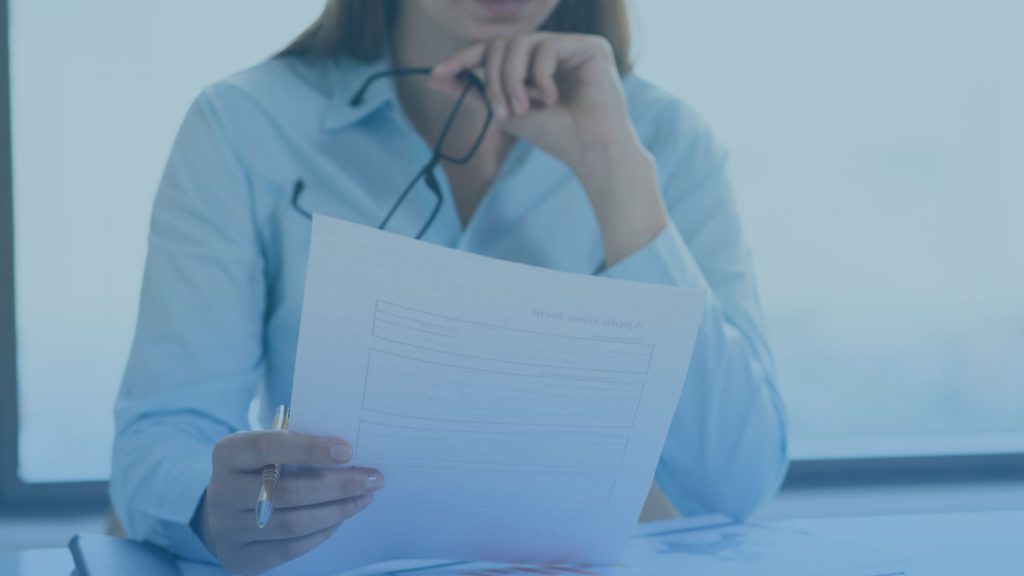 Mulher branca segurando um óculos e olhando para um documento. A imagem representa uma profissional analisando os dados preenchidos na guia TISS.