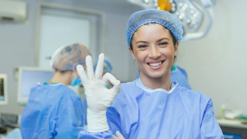 Mulher vestida com jaleco de cirurgia fazendo sinal de ok. A imagem representa que ela é uma auxiliar, e posteriormente haverá a cobrança por honorários de auxiliares de cirurgia.