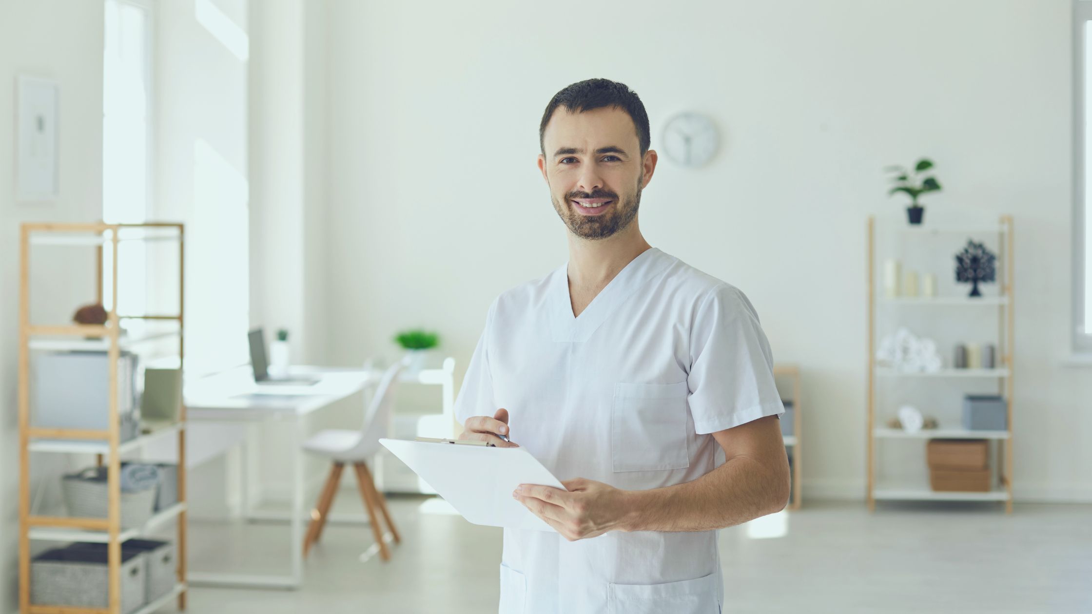 Homem sorrindo segurando prancheta em uma sala de fisioterapia. Imagem simboliza que ele é o profissional que posteriormente fará o faturamento da fisioterapia..