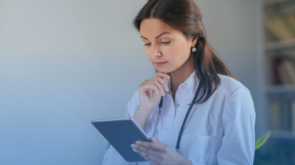 Mulher vestida com um jaleco de médico. Ela está com uma das mãos no queixo e a outra segurando um tablet. A imagem representa uma profissional de saúde analisando os modelos de remuneração em diárias hospitalares.